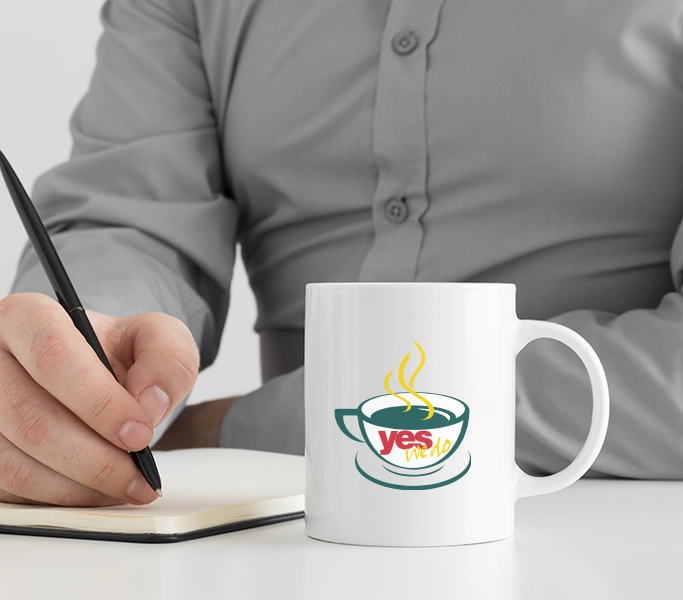 A man writing notes with a coffee cup in front of him with Yes We Do Coffee logo on the cup